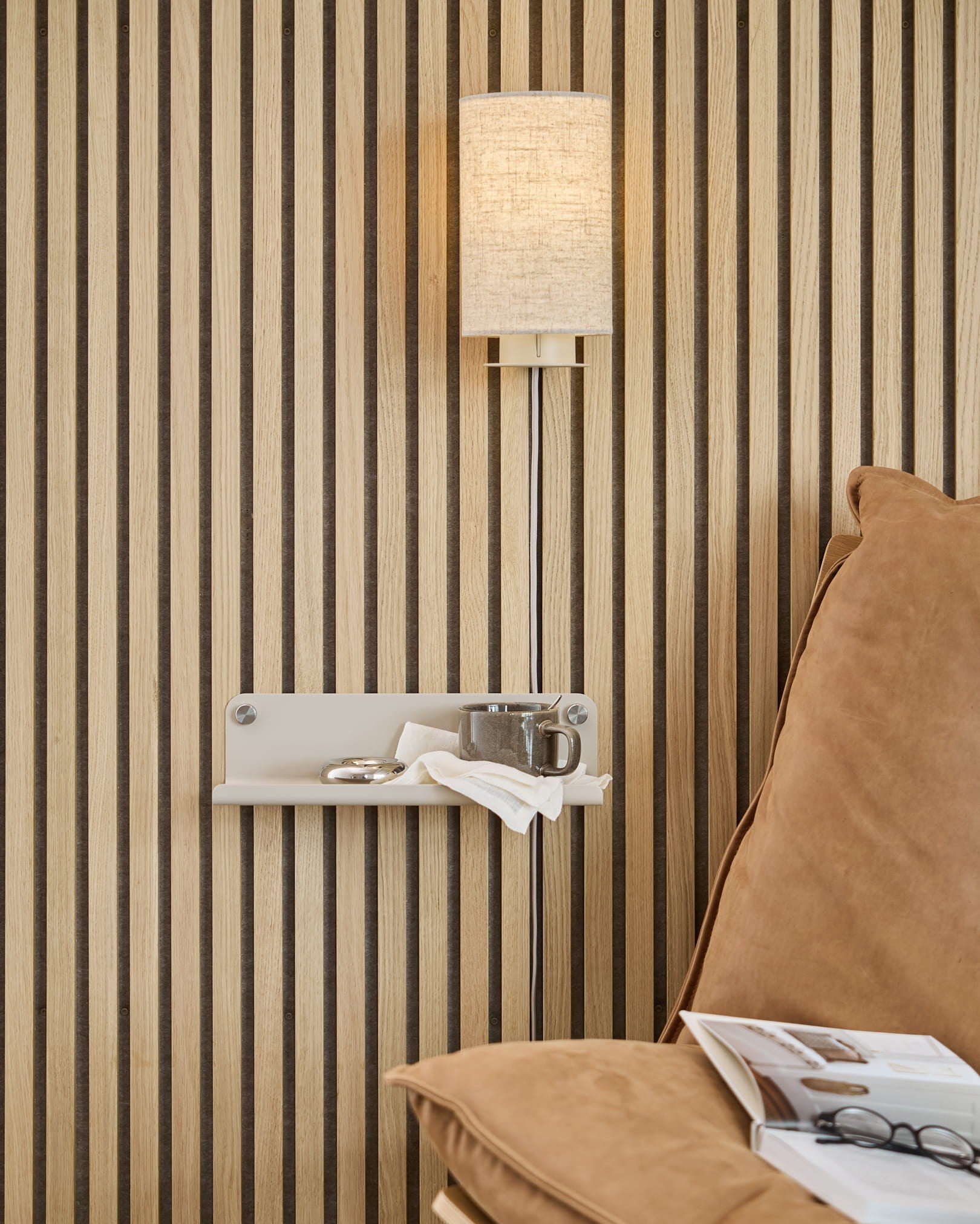 Mur acoustique à lattes de bois « Akupanel » disposées verticalement, applique murale beige, petite étagère sur laquelle sont posés une tasse et une serviette, canapé beige sur lequel est posé un magazine ouvert.